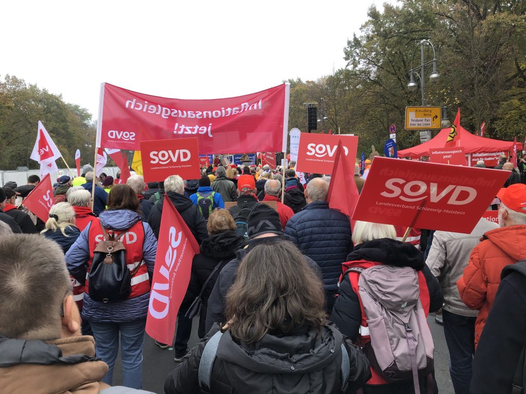 Menschen dicht aneinander stehend, von hinten zu sehen. Viele rote Plakate mit SOVD Landesverband Schleswig-Holstein. Ein Transparent "Infltionsausgleich..." Links vorne eine ver.di-Fahne. Rechts ein Pavillon, darüber gelbes Verkehrsschild. Unter dem Transparent hindurch ist die Bühne mit Redner zu sehen.