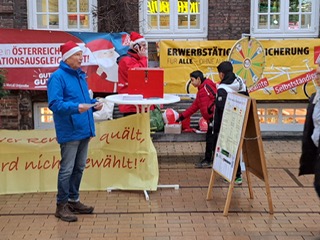 Transparente, und Stelltafel vor einer Hauswand. Eine Person mit roter Mütze vorne, zwei weitere im Hintergrund in Aktion.