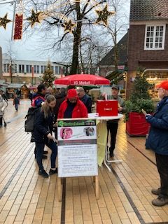 An einer Stelltafel stehen Leute zum lesen. Die Pflasterung der Umgebung glänzt regennass. Auf einem Tisch eine rote "Wahlurne". Weihnachtliche Sterne hängen in den Bäumen.