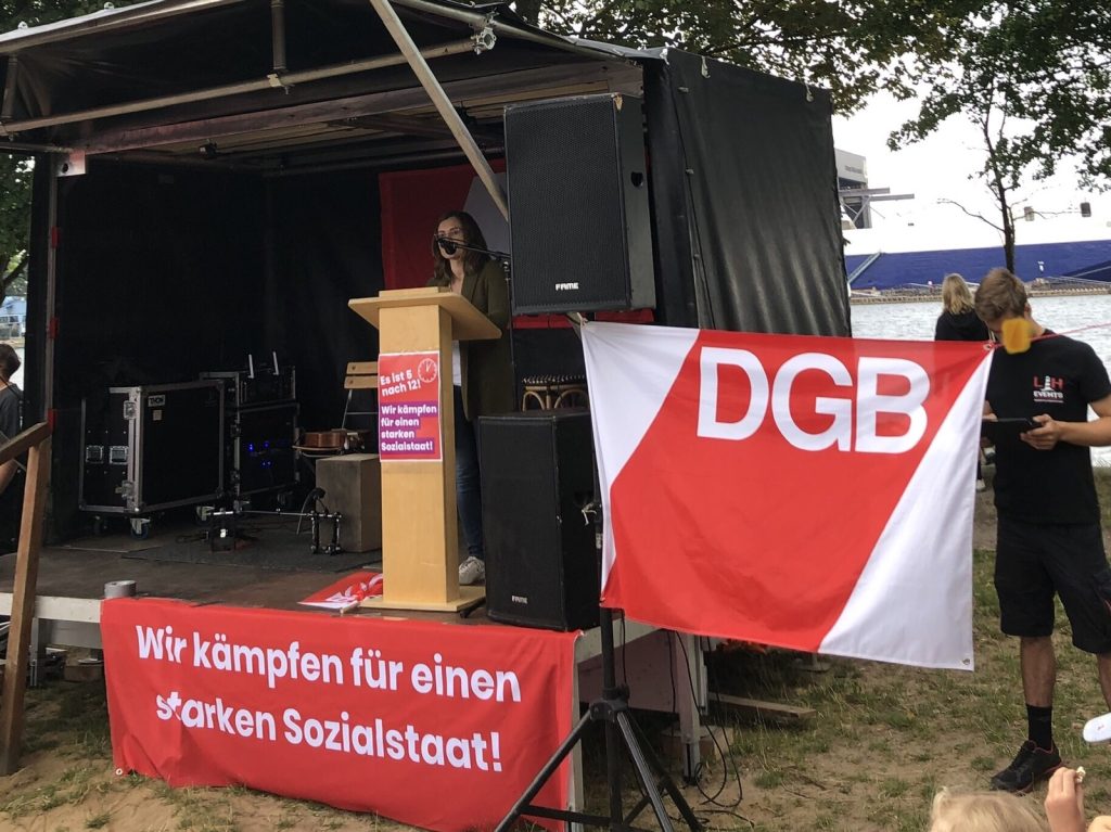 Eine Bühne mit redepult an der eine Person steht. Ein Plakat mit dem Motto hängt am Pult. Ein Transparent "Wir kämpfen für einen starken Sozialstaat hängt unten. Daneben eine DGB-Fahne. Im Hintergrund die Förde.